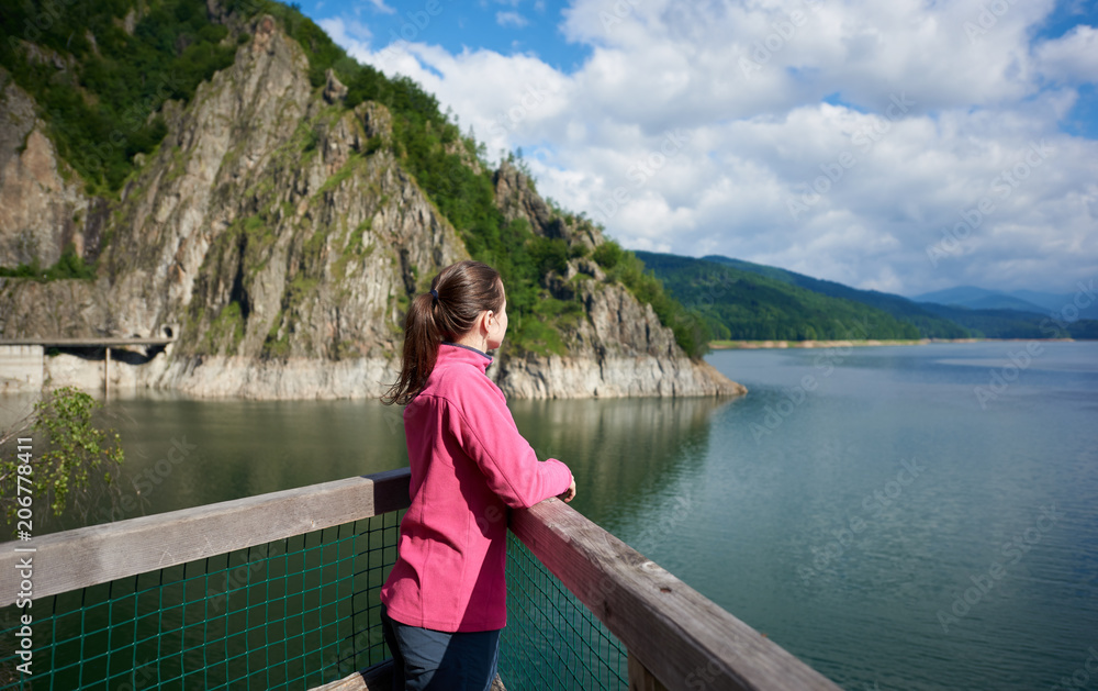 Brunette female tourist looking far into the distance admiring magnificent views of green rocky mountains and breathtaking Vidraru lake in Romania. Woman amazing beautiful scenery landscape happy calm