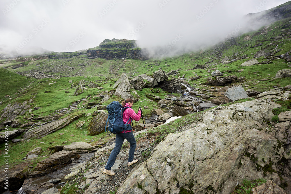 Girl hiker climbs to the top of the mountainous country of Romania. Clouds shroud mountains. Woman with backpack and trekking sticks