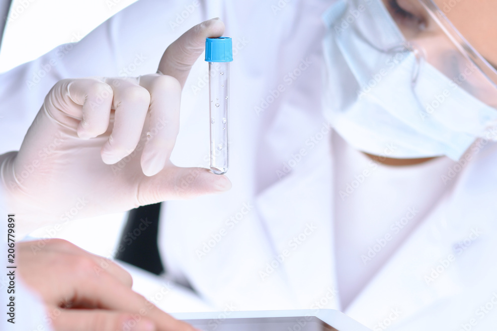 Closeup of scientific research team with clear solution in laboratory. Blonde female chemist holds test tube of glass while her colleague checks results with tablet pc. Blood test, medicine or Closeup of scientific research team with clear solution in laboratory. Blonde female chemist holds test tube of glass while her colleague checks results with tablet pc. Blood test, medicine or