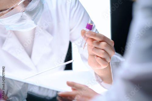 Closeup of scientific research team with clear solution in laboratory. Blonde female chemist holds test tube of glass while her colleague checks results with tablet pc. Blood test, medicine or