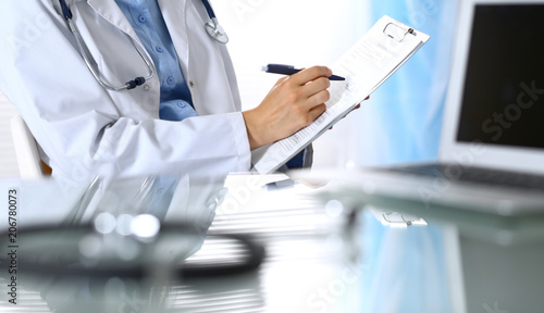 Female doctor filling up medical form on clipboard, closeup. Reflecting glass table is a physician working place. Healthcare, insurance and medicine concept