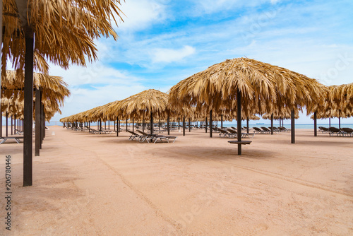 Beach straw umbrellas or palapas and sunbeds on the sandy deserted sea shore. Cargo ship is on far background. Cloudy blue sky.