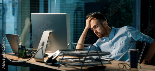 Bild auf Leinwand Young tired guy leaning on hand looking in monitor of computer and brainstorming while working late alone