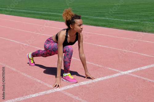 A young beautiful dark-skinned girl in a sporty black T-shirt and pink sneakers is preparing to race in the stadium starting line next to the green grass. Life style. Copy space.