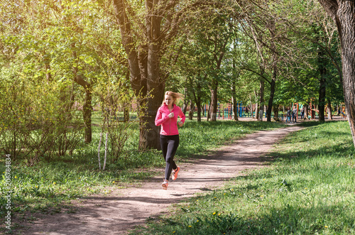 A young athletic girl in a pink and black sports suit jogging along the path in the park in the morning. © Olga