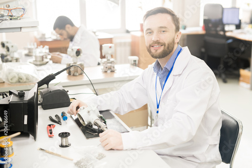 Cheerful confident bearded male repairing engineer in lab coat sitting at table with tools and dissembled measuring device and looking at camera