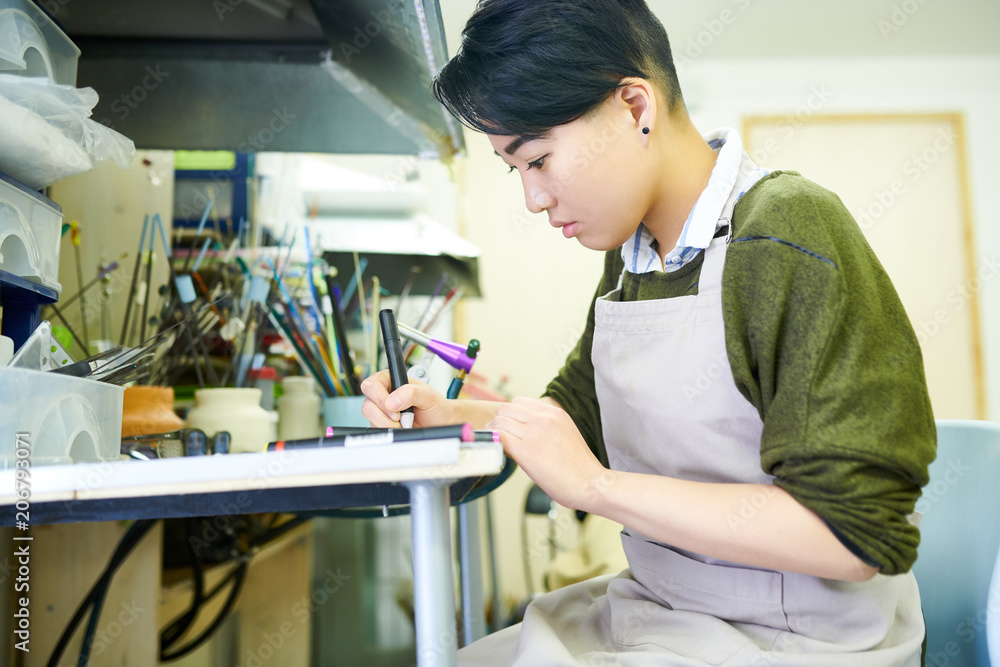 Side view portrait of young Asian woman drawing sketches in glassworking workshop, ready to create beautiful jewelry Side view portrait of young Asian woman drawing sketches in glassworking workshop, ready to create beautiful jewelry
