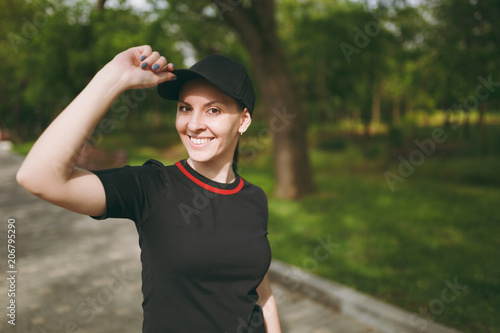 Young athletic smiling beautiful brunette girl in black uniform and cap standing, looking on camera keeping hand near cap on training on path in city park outdoors. Fitness, healthy lifestyle concept.