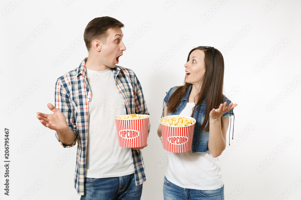 Young shocked couple, woman and man in 3d glasses and casual clothes watching movie film on date, holding buckets of popcorn, spreading hands isolated on white background. Emotions in cinema concept.