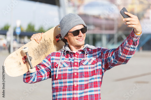 Portrait of hipster man with sunglasses dressed in cheked shirt and cap holding skateboard and making selfie on smartphone photo