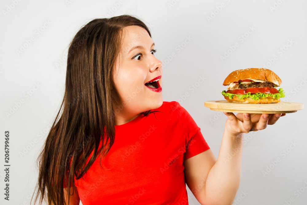 Young fat girl looks at the burger and smiles on gray background.