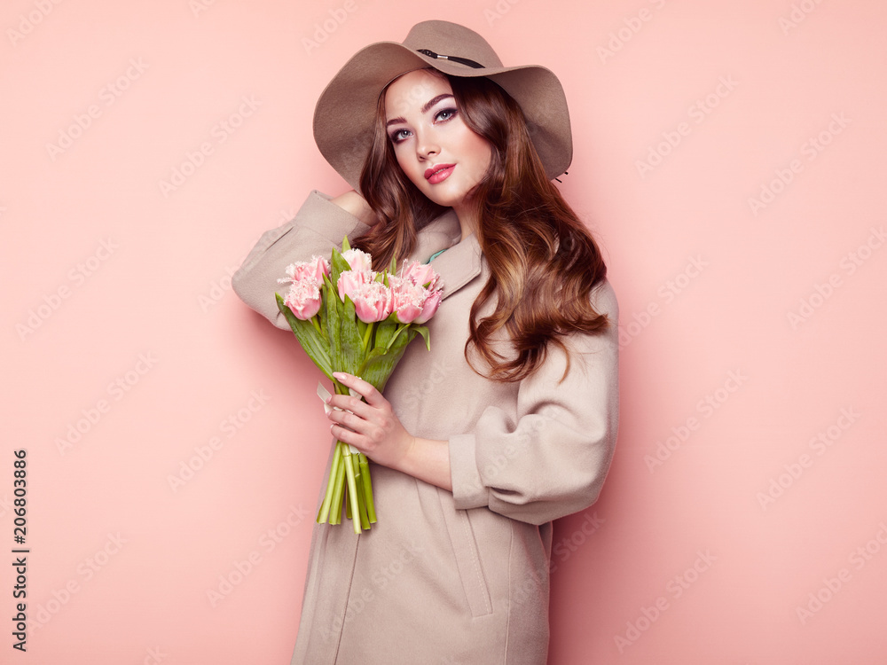 Brunette Young Woman in Stylish Greatcoat and Hat. Girl in Beige Coat with Bouquet of Flowers. Model with Long Curly Hair. Spring Lookbook Brunette Young Woman in Stylish Greatcoat and Hat. Girl in Beige Coat with Bouquet of Flowers. Model with Long Curly Hair. Spring Lookbook