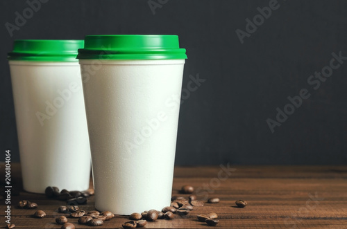 two white plastic coffee cups with a green lid on a wooden table with scattered coffee beans  front view with copy space