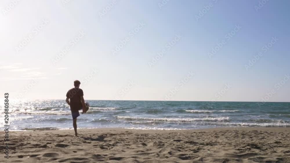 Young man in swimming trunks does tricks dribbling bouncing football on the sea shore at the beach at sunset silhouette wide shot Young man in swimming trunks does tricks dribbling bouncing football on the sea shore at the beach at sunset silhouette wide shot