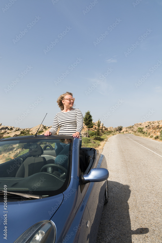 Young blonde woman driving in convertible blue rental car without roof on mountain road in Naxos island, Greece