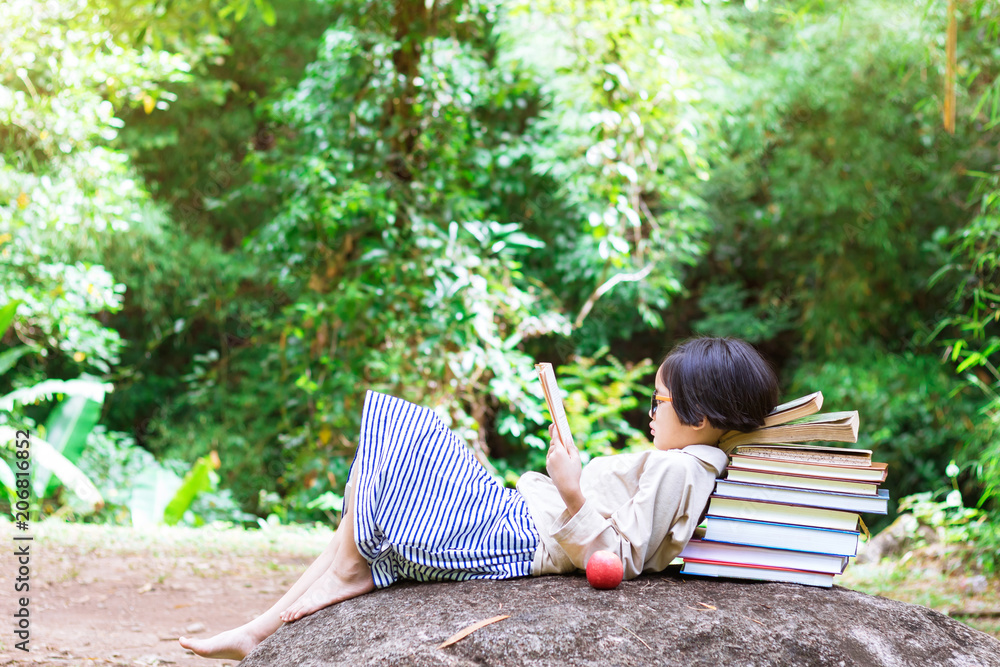 Fototapeta premium Asian little girl with glasses lying on stone reading a books
