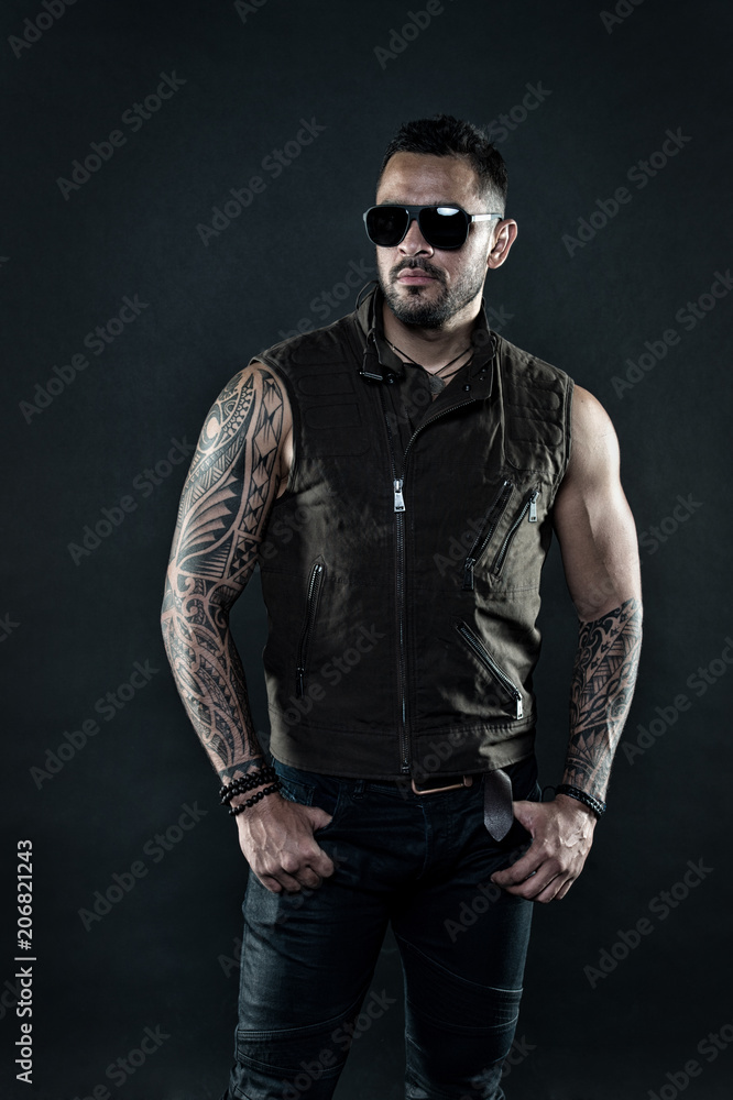 Fashion macho in trendy sunglasses. Tattoo model with beard on unshaven  face. Bearded man with tattoo on strong arms. Tattooed man with biceps and  triceps. Fashion style and trend, vintage filter Stock