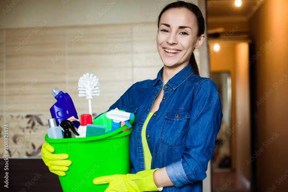 Beautiful young woman in protective gloves is holding a green bucket with equipment for cleaning. Girl looking at camera and smiling Beautiful young woman in protective gloves is holding a green bucket with equipment for cleaning. Girl looking at camera and smiling