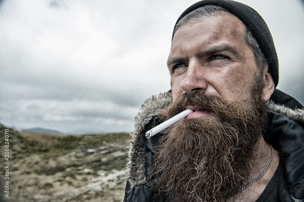 Fotka „Man with long beard and mustache smoking cigaret. Brutality concept.  Hipster on strict face with beard looks brutally while hiking and smoking.  Man with brutal bearded appearance, untidy and unshaven“ ze