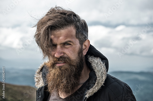 Man with brutal bearded appearance, brutal unshaven man looks untidy. Man with long beard and mustache wears jacket. Hipster on strict face with beard looks brutally while hiking. Hermit concept photo