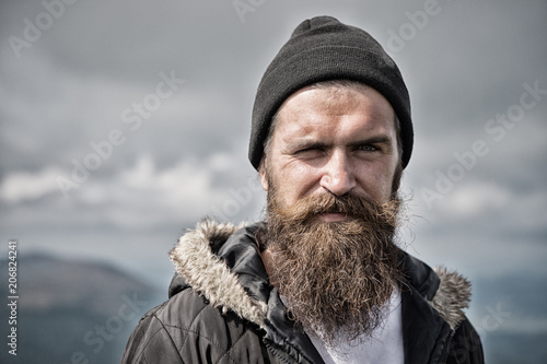 Man with long beard and mustache wears hat. Hipster on strict face with beard looks brutally while hiking. Masculinity concept. Man with brutal bearded appearance, brutal unshaven man looks untidy