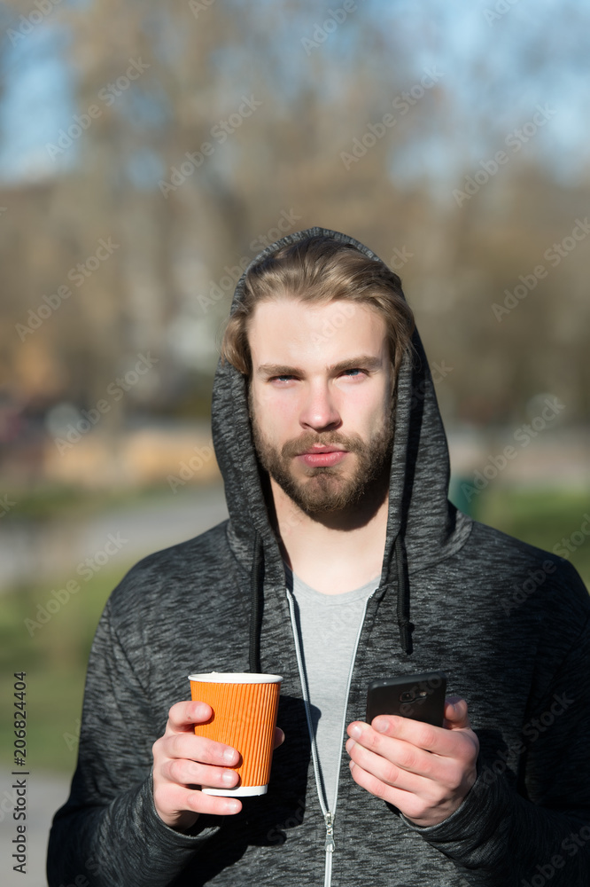 Guy with smartphone and takeaway drink on fresh air. Bearded man in hood hold disposable coffee cup on sunny outdoor. Coffee or tea mood. Morning tea for breakfast. New technology and modern life Guy with smartphone and takeaway drink on fresh air. Bearded man in hood hold disposable coffee cup on sunny outdoor. Coffee or tea mood. Morning tea for breakfast. New technology and modern life