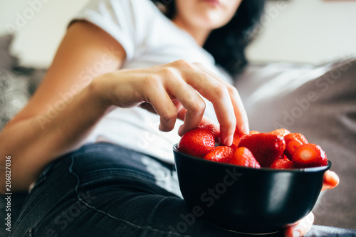 Woman sitting on couch and eating strawberries out of bowl. Healthy eating, vegetarian food, fruit. photo