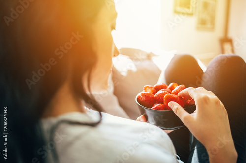 Woman sitting on couch and eating strawberries out of bowl. Healthy eating, vegetarian food, fruit.