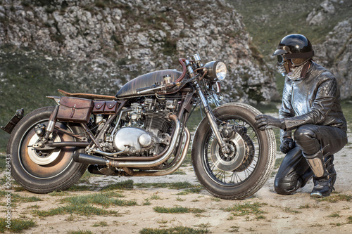 Biker with black leather suit and mask stay rit on the right of his custom special rat motorbike. Desolated rocks in the background. Post apocalyptic concept