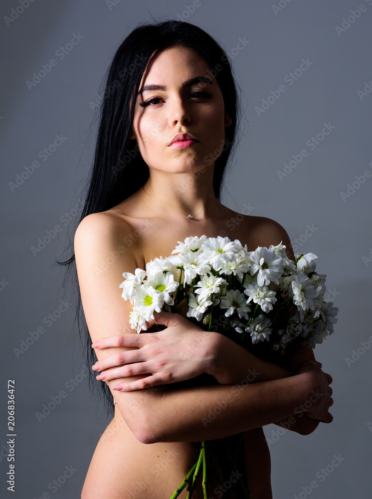 Woman with smooth healthy skin looks attractive. Natural cosmetics concept. Girl on calm face naked holds chamomile flowers in front of breasts. Lady covers breasts with flowers, grey background. Woman with smooth healthy skin looks attractive. Natural cosmetics concept. Girl on calm face naked holds chamomile flowers in front of breasts. Lady covers breasts with flowers, grey background.