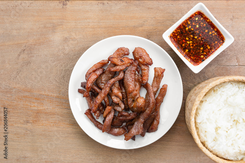 top view of fried sun dried pork in a ceramic dish served with spicy sauce and sticky rice on wooden table. thai style delicious food menu.