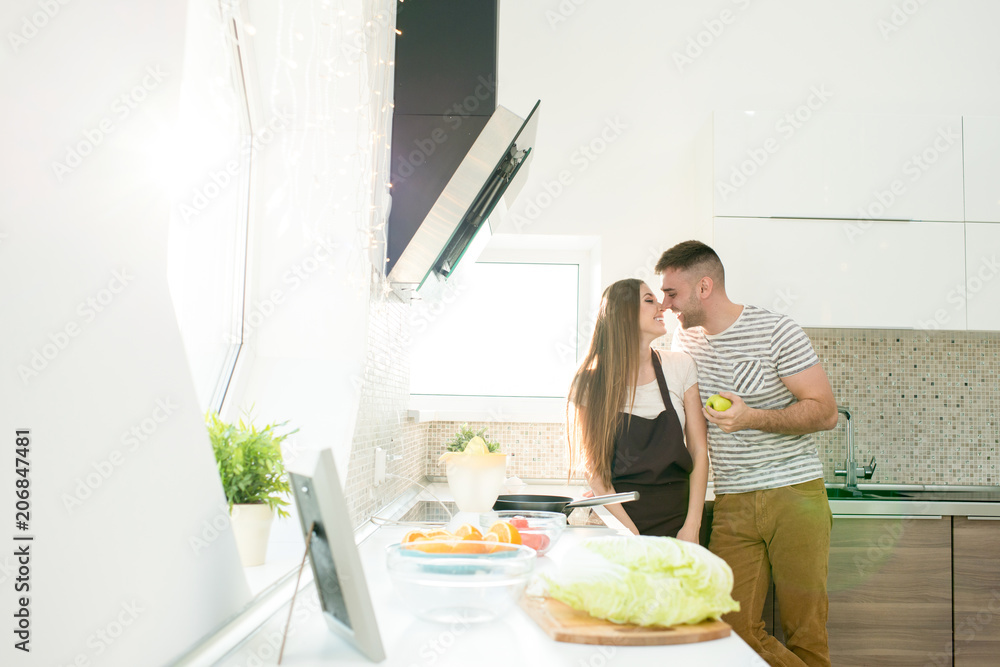 Cheerful happy young couple in casual clothing enjoying time together in kitchen: handsome man with apple reaching to kiss wife in apron