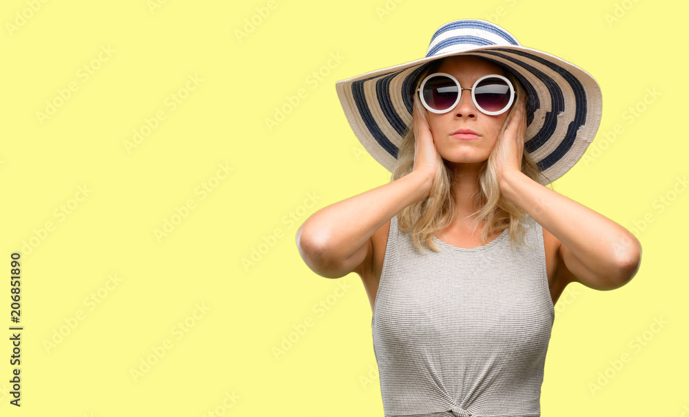 Young woman wearing sunglasses and summer hat covering ears ignoring annoying loud noise, plugs ears to avoid hearing sound. Noisy music is a problem. Young woman wearing sunglasses and summer hat covering ears ignoring annoying loud noise, plugs ears to avoid hearing sound. Noisy music is a problem.