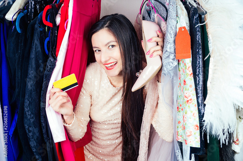 beautiful crazy brunette mad woman shopaholic looks through the hangers wardrobe with clothes holding a shoes and a plastic credit card in her hand . concept of seasonal sales and shopaholicism photo