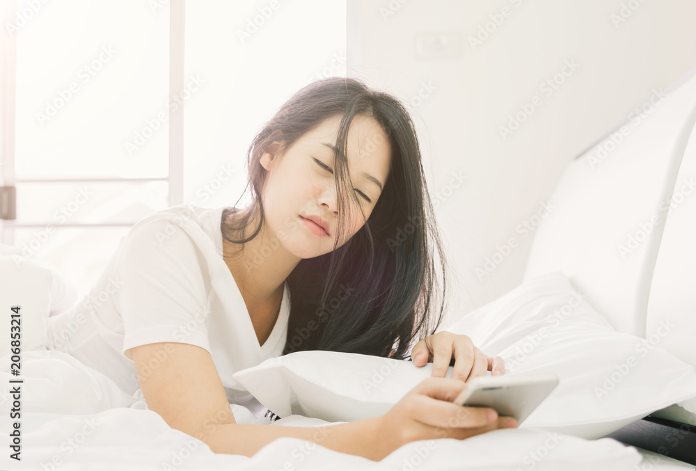 Young asian beautiful woman hates waking up early in the morning. Sleepy girl looking at alarm clock with smartphone Young asian beautiful woman hates waking up early in the morning. Sleepy girl looking at alarm clock with smartphone