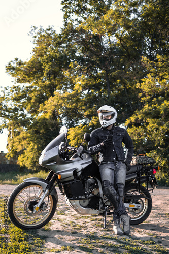 Fototapeta Naklejka Na Ścianę i Meble -  rider guy use smart phone in jeans biker jacket and helmet sit on tourist touring motorcycle. outdoors, dual sport adventure concept, vertical photo, dirt road