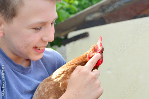 The boy holds a brown chicken in his hands and smiles and kisses her and hugs and holds a finger for his beak