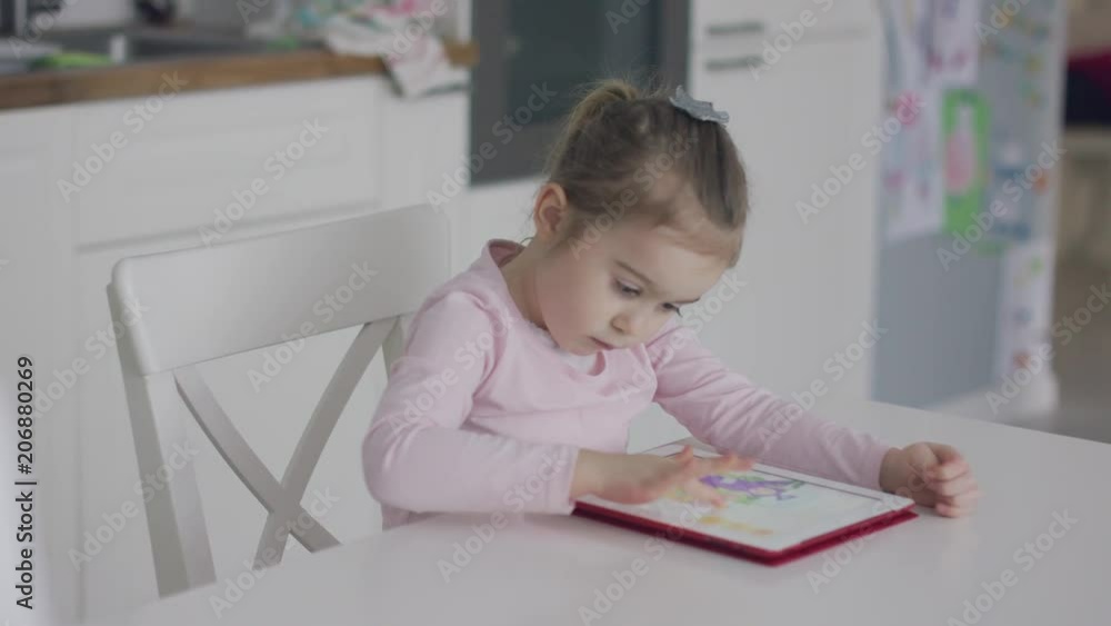 Little adorable girl sitting at table in kitchen and tapping on touchscreen of tablet while playing game alone and looking focused. Little adorable girl sitting at table in kitchen and tapping on touchscreen of tablet while playing game alone and looking focused.