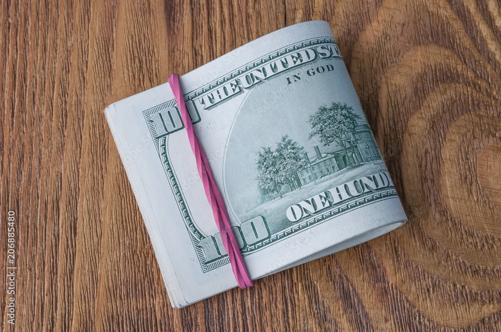 A pack of one hundred dollar bills stretched by an elastic band on a wooden table. A pack of one hundred dollar bills stretched by an elastic band on a wooden table.