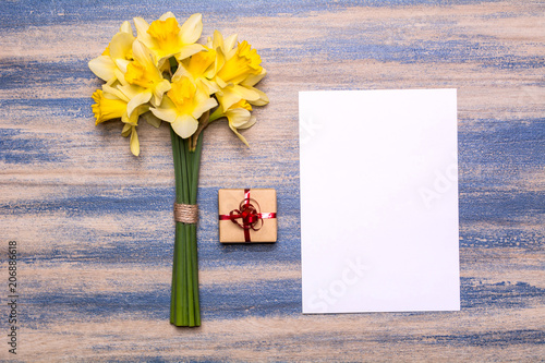 Fototapeta Naklejka Na Ścianę i Meble -  Workplace with a gift with a red ribbon, a piece of paper and a bouquet of daffodils on a blue wooden background. Flat lay design, top view.