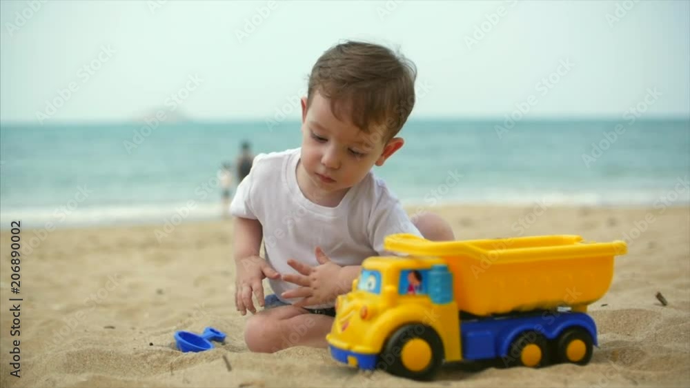 Two-year old beautiful baby toddler boy riding a toy cargo car on summer beach closeup. Slow motion Two-year old beautiful baby toddler boy riding a toy cargo car on summer beach closeup. Slow motion