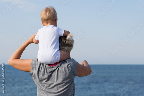 Little baby in white shirt sits on shoulders of his mother watching blue sea. View from behind. Mom in grey shirt pointing forward. Vacation with childrend and mom and son relationship concept photo