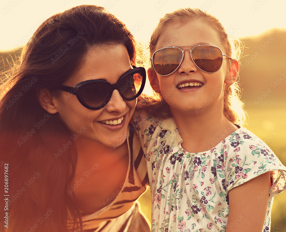 Obraz premium Happy fashion kid girl embracing her mother in trendy sunglasses and looking on nature background. Closeup toned color portrait of happiness.
