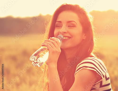 Happy smiling woman drinking water from the bottle on summer bright outdoor background. Closeup toned orange color portrait