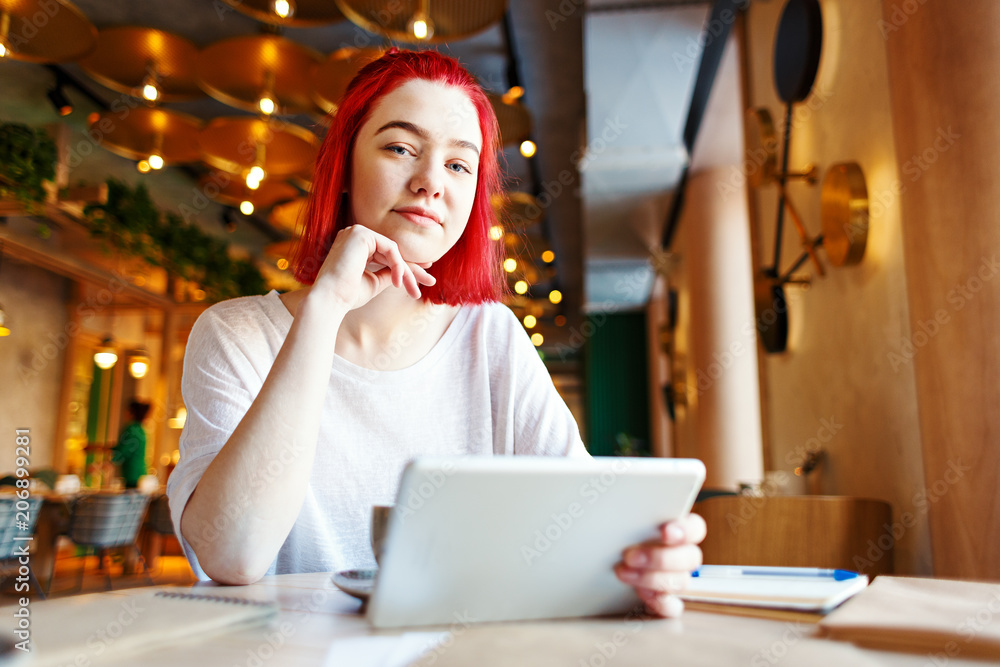 Portrait of girl with red hair looking at camera while sitting at cafe table with tablet computer in her hands Portrait of girl with red hair looking at camera while sitting at cafe table with tablet computer in her hands