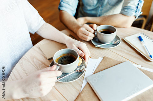 Hands of unrecognizable man and woman having coffee during business meeting in cafe, tablet computer, notepad and papers on table nearby