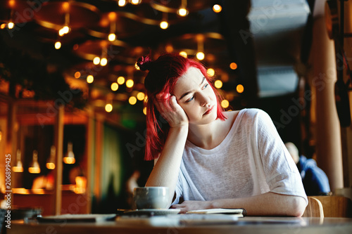 Portrait of beautiful young woman with red hair sitting alone at table in cafe. Girl looking sadly through window and waiting for somebody to come