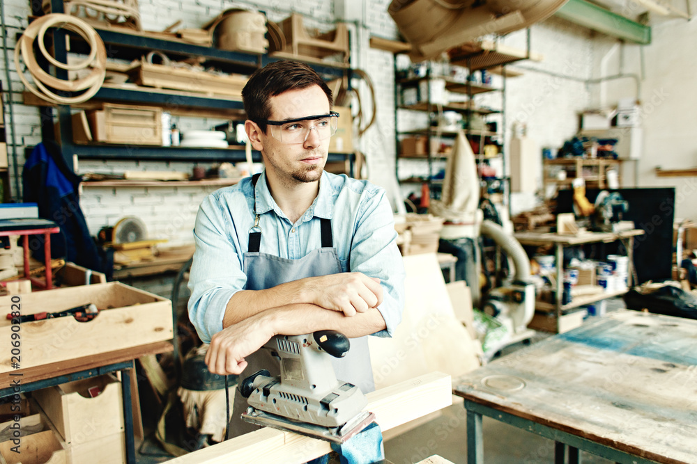 Portrait of thoughtful woodworker in protective eyewear leaning on sanding machine while resting from work in his workshop Portrait of thoughtful woodworker in protective eyewear leaning on sanding machine while resting from work in his workshop