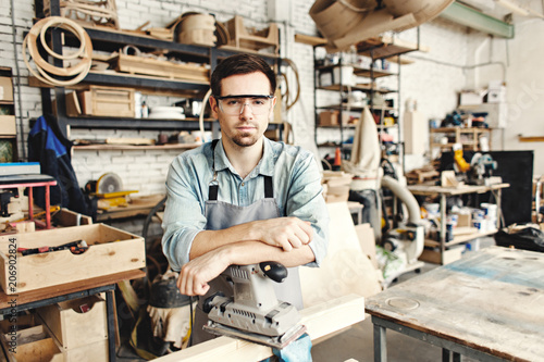Portrait of professional young carpenter posing at workplace leaning on sanding machine. He looking at camera through protective eyewear