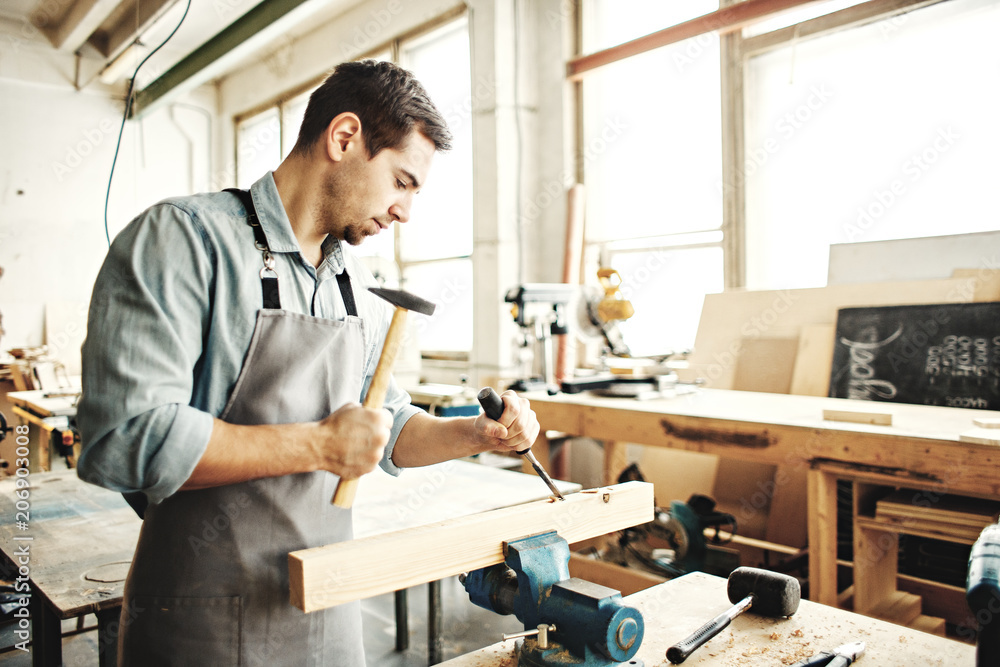Young carpenter working in his shop, using hammer and wood chisel to make mortise in piece of wood Young carpenter working in his shop, using hammer and wood chisel to make mortise in piece of wood